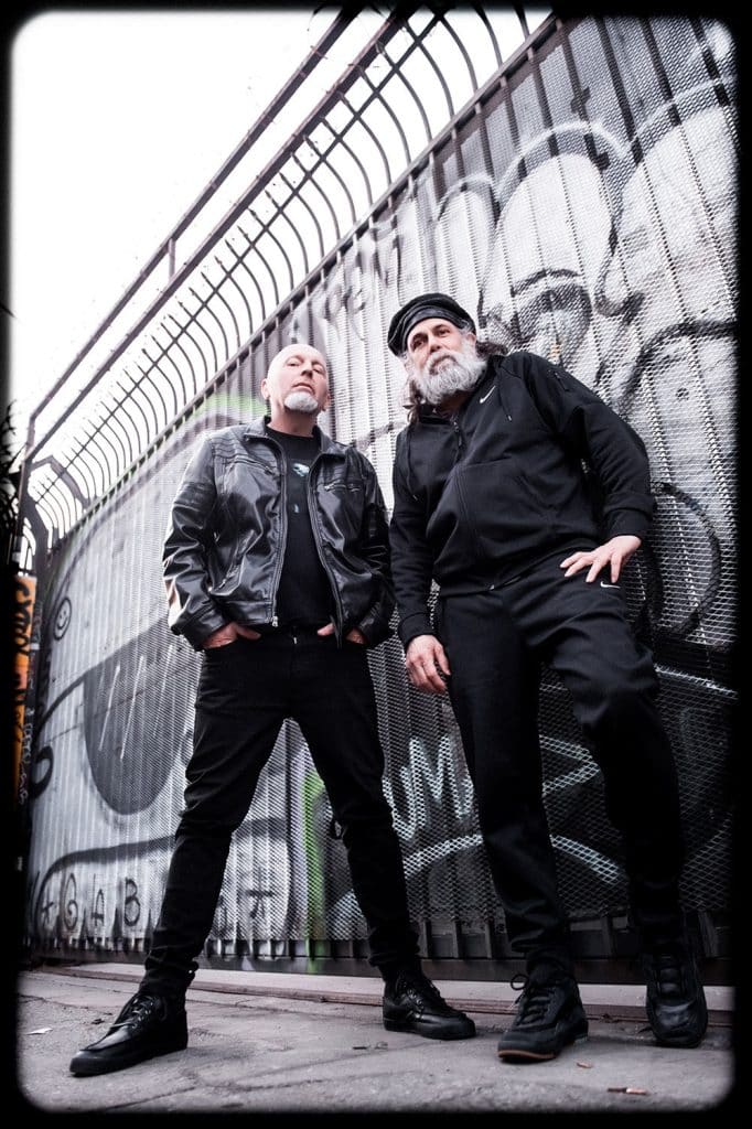 Chris Barnes and Jack Owen of Six Feet Under pose in front of a graffiti-covered metal fence. Both wear all black streetwear, exuding a gritty urban metal aesthetic ahead of the Midnight In Hell 2025 tour.