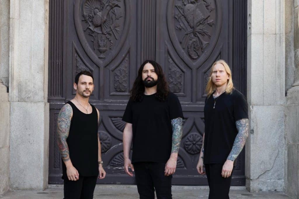 Three members of Born of Osiris stand in front of a large, ornate, dark wooden door with carved religious and floral imagery. All wear black clothing and display tattooed arms. The central figure has long hair and a beard, flanked by two members with shorter hair, one with a sleeveless shirt and the other with long blond hair.