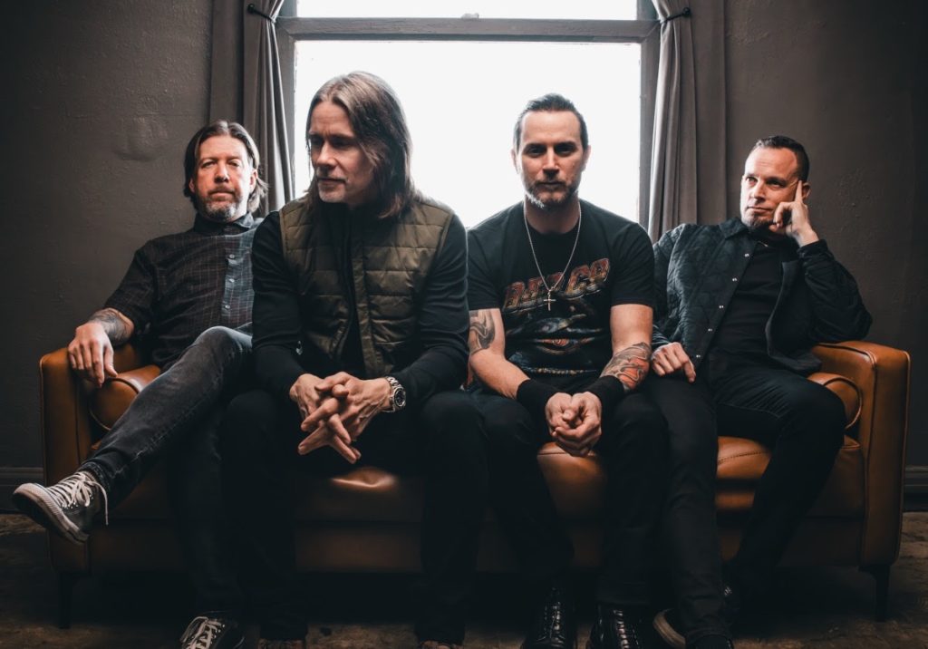 Alter Bridge band members seated on a leather couch in a dimly lit room, featuring Brian Marshall, Myles Kennedy, Scott Phillips, and Mark Tremonti, promoting their 2026 self-titled album