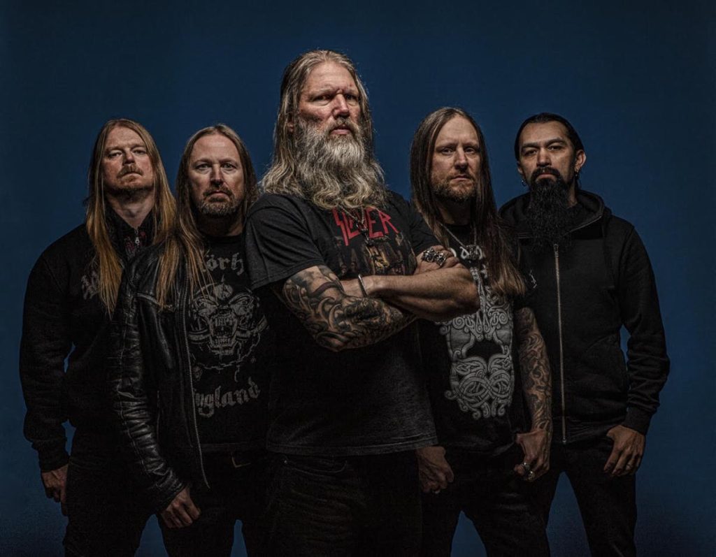Amon Amarth band photo featuring all five members standing against a dark blue backdrop. Johan Hegg stands front and center with arms crossed, wearing a black Slayer shirt, flanked by bandmates with long hair and dark clothing, exuding a powerful and stoic Viking metal presence