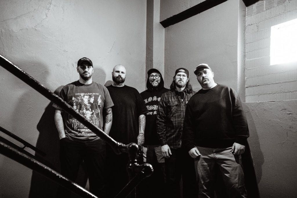 Black and white promo photo of The Acacia Strain featuring all five band members standing against a concrete wall and staircase, wearing casual streetwear and band merch, lit dramatically from the side.
