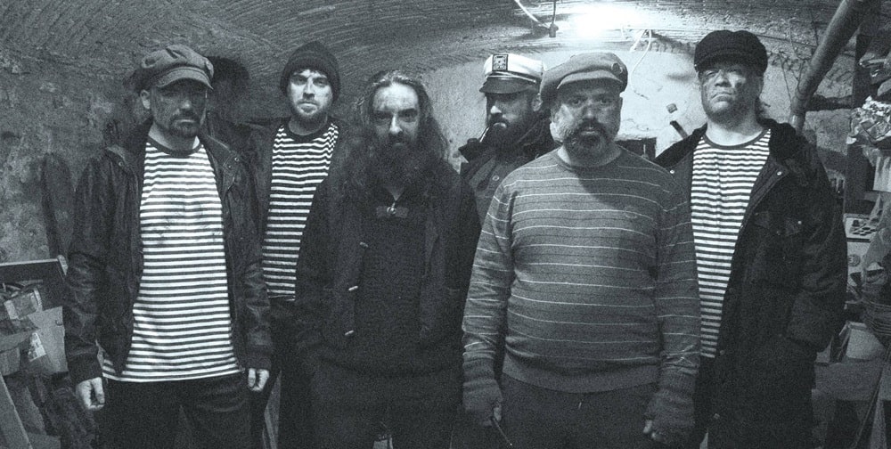 Calico Jack band members dressed in pirate-themed attire, standing in a dimly lit stone cellar with arched ceiling, wearing striped shirts and captain hats, evoking a rugged maritime aesthetic.