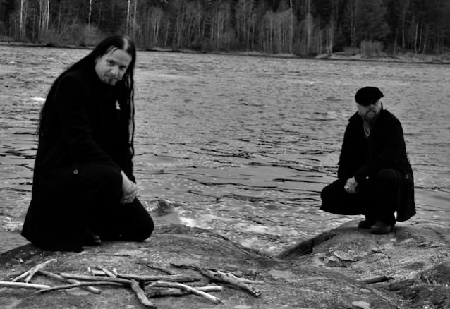 DIMMU BORGIR members Shagrath and Silenoz pose by a river in black attire, surrounded by nature. Black and white photo emphasizing the band’s dark aesthetic.
