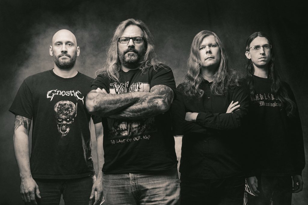 Gorguts band photo featuring four members standing in a dark setting, arms crossed, wearing black shirts with death metal logos.