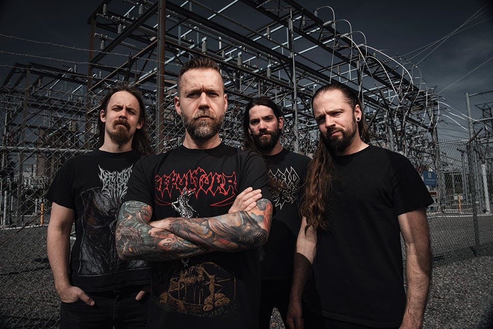 Revocation band members standing in front of a fenced electrical substation, wearing black metal shirts, with vocalist/guitarist Dave Davidson at the front crossing his tattooed arms.