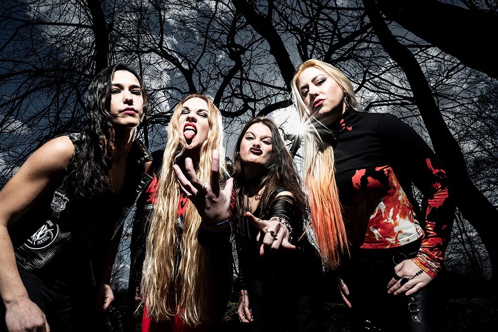Brazilian thrash metal band Nervosa posing outdoors under bare trees, with one member snarling at the camera and flashing the horns hand gesture.