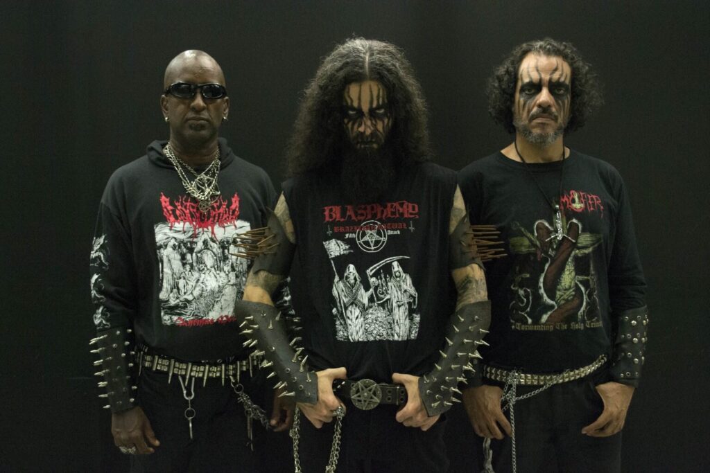 Brazilian black metal band Mystifier standing in a dark studio, wearing corpse paint, spikes, and studded belts.