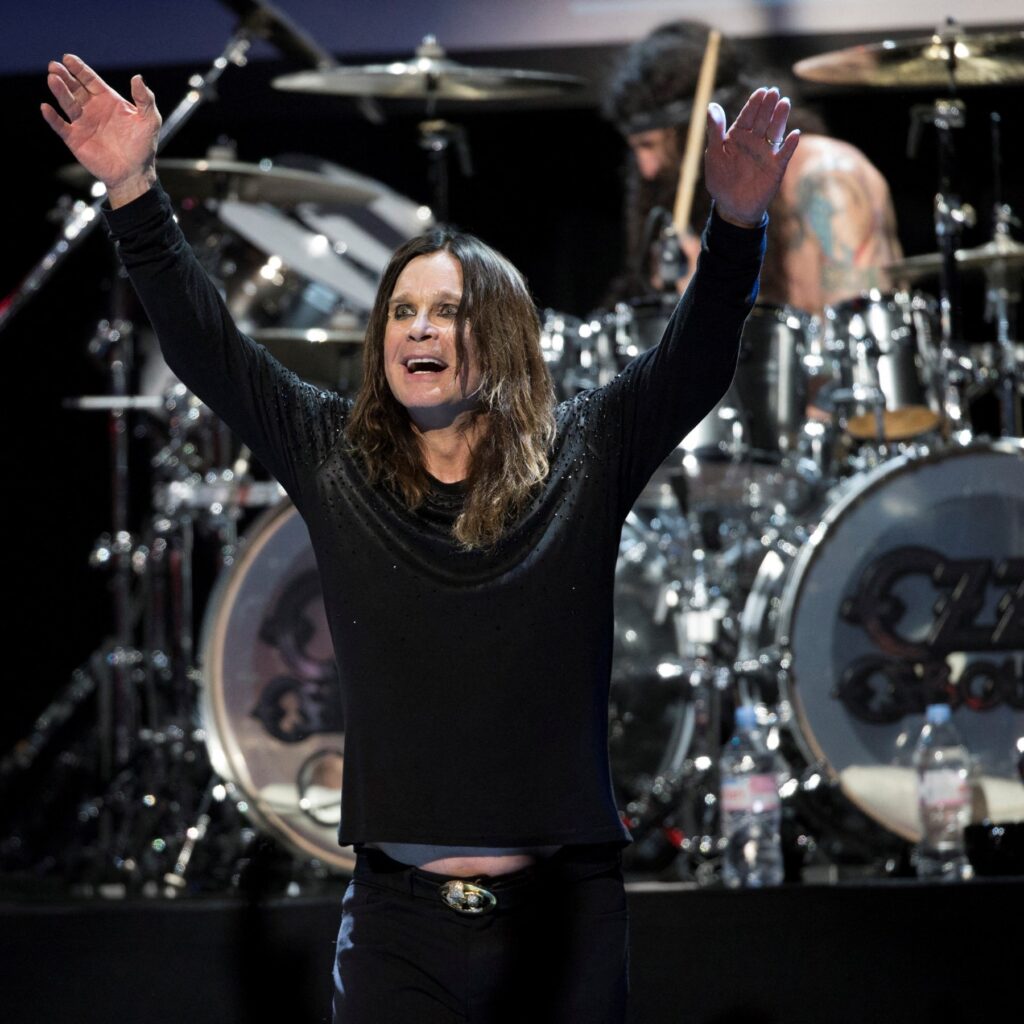 Ozzy Osbourne onstage with arms raised, smiling as he stands in front of a drum kit during a live performance.