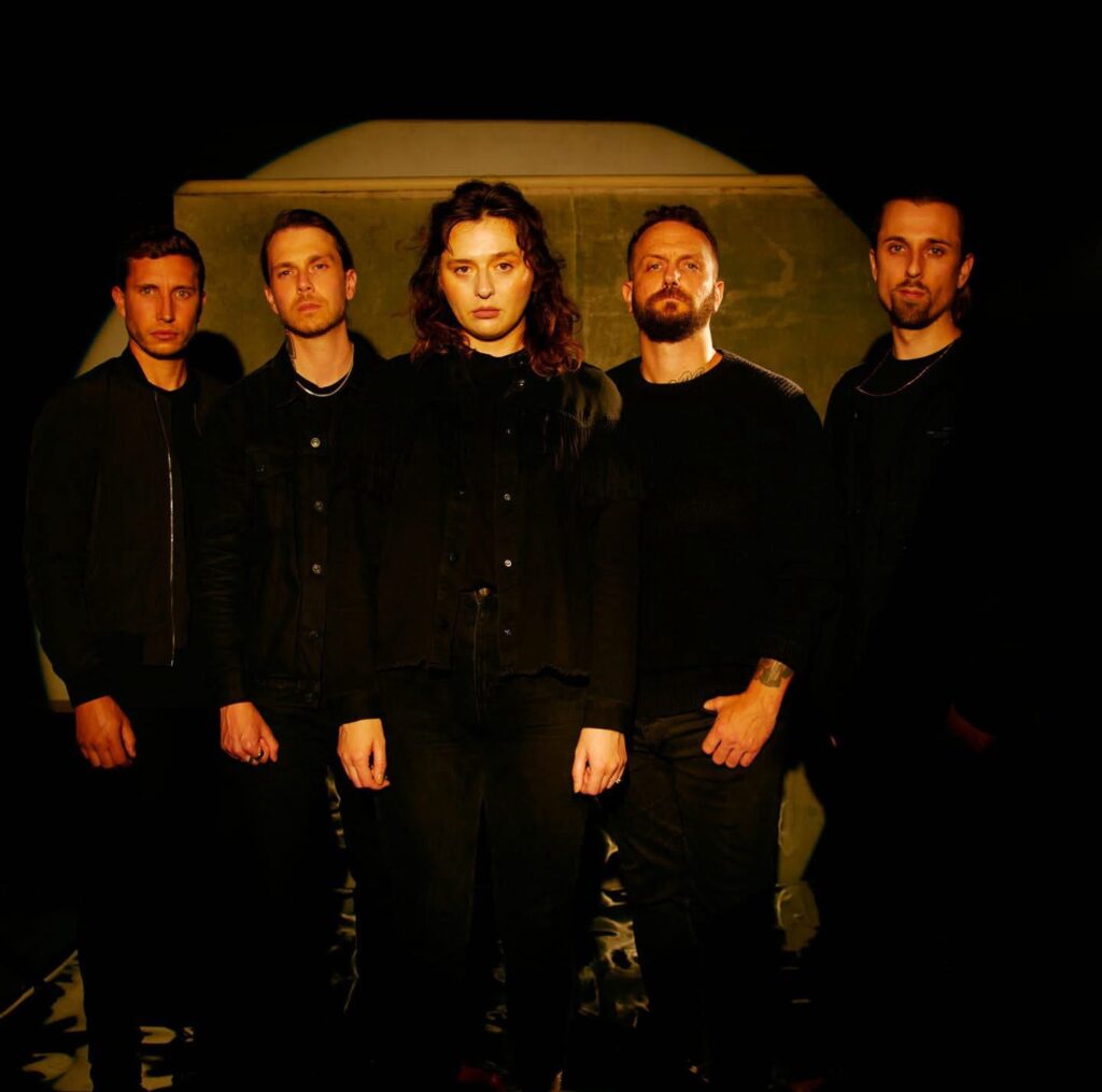“Venom Prison band portrait. Five members standing in a dim, golden lit space, all dressed in black, facing the camera with a serious expression.”