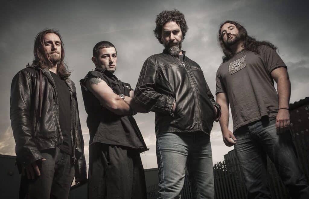 “Hunted band photo with four members standing outdoors under a moody sky, wearing dark casual clothing and leather jackets, looking toward the camera.”