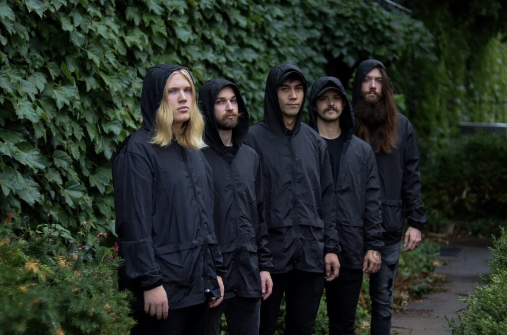 Band photo of Ben’s Raincoat: five members standing outdoors in matching black raincoats, surrounded by ivy covered walls and greenery.