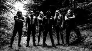 Hecate Enthroned standing in a dark forest in black and white promotional photo surrounded by towering trees and shadowed atmosphere.