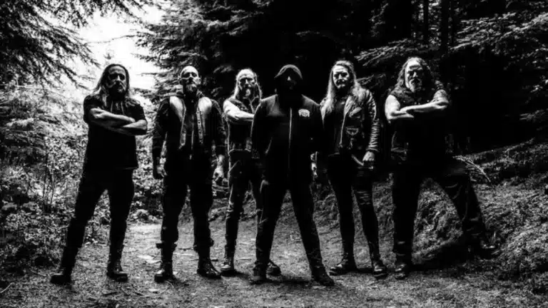 Hecate Enthroned standing in a dark forest in black and white promotional photo surrounded by towering trees and shadowed atmosphere.