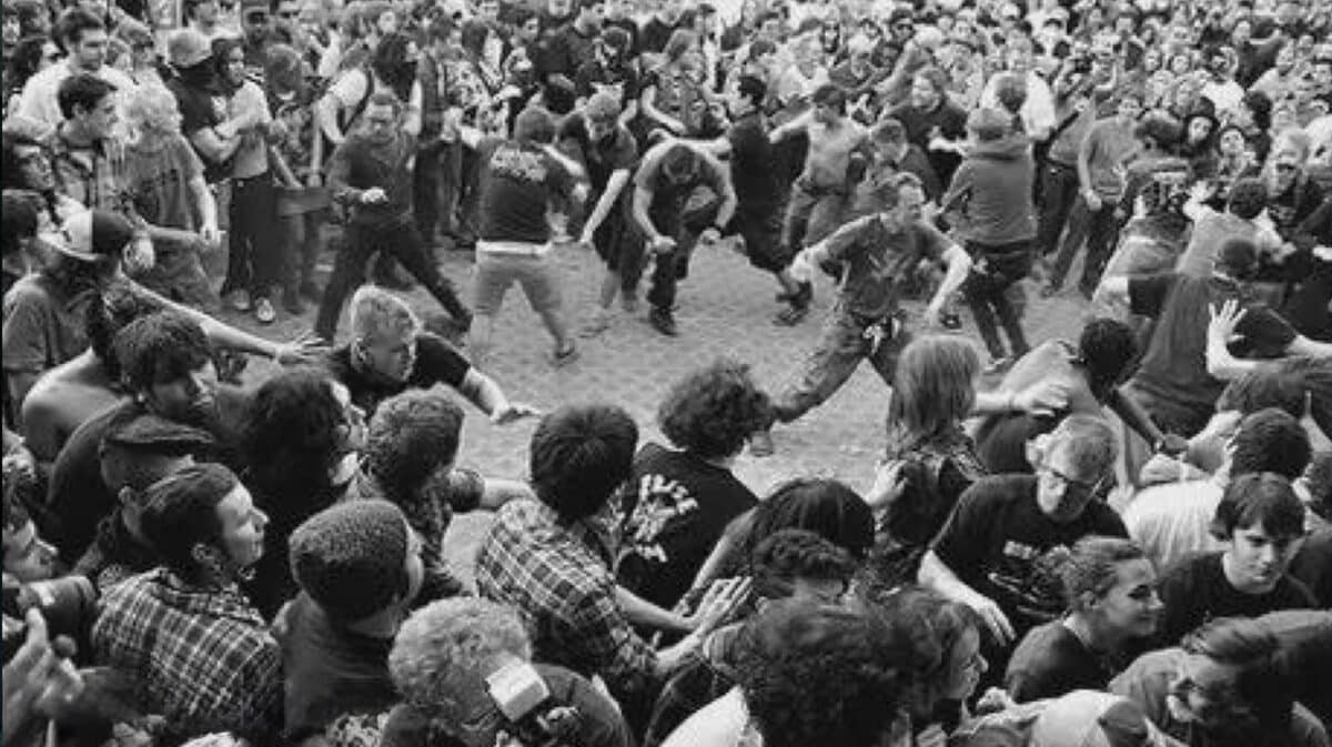 Black-and-white photo of a large circle pit at a heavy metal concert with fans running, moshing, and surrounding the open pit during a live performance.