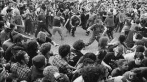 Black-and-white photo of a large circle pit at a heavy metal concert with fans running, moshing, and surrounding the open pit during a live performance.