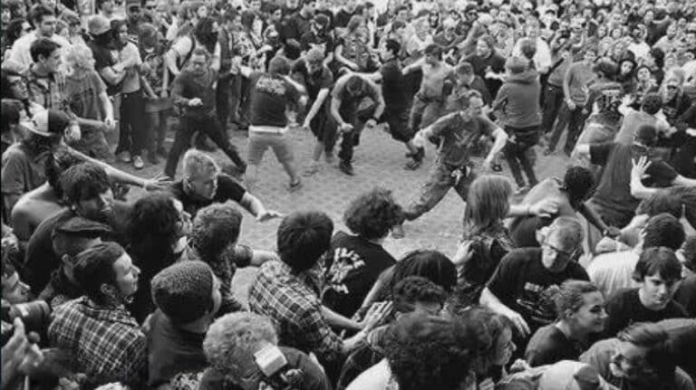 Black-and-white photo of a large circle pit at a heavy metal concert with fans running, moshing, and surrounding the open pit during a live performance.