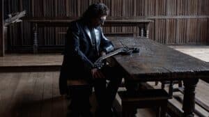Tony Iommi seated beside a wooden table with his signature guitar in a promotional photo by Ross Halfin.