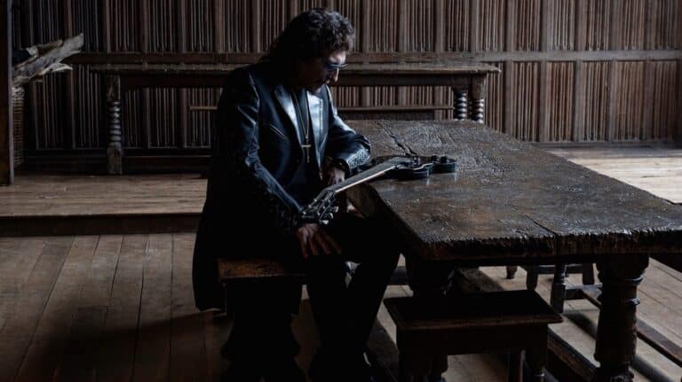 Tony Iommi seated beside a wooden table with his signature guitar in a promotional photo by Ross Halfin.