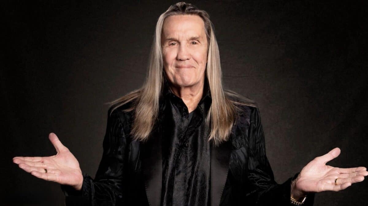 Nicko McBrain Donald Trump interview: Iron Maiden drummer poses with both hands raised, wearing a black jacket against a dark background.