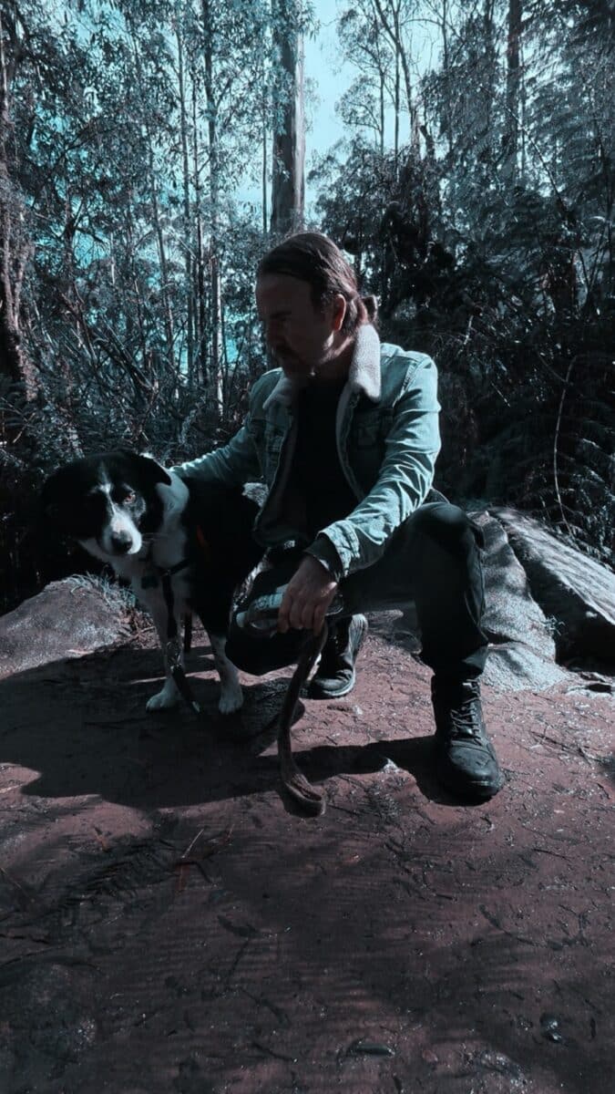Craig Bannerman Interview featured photograph of the Australian extreme metal musician crouching beside his black-and-white dog in a wooded landscape.